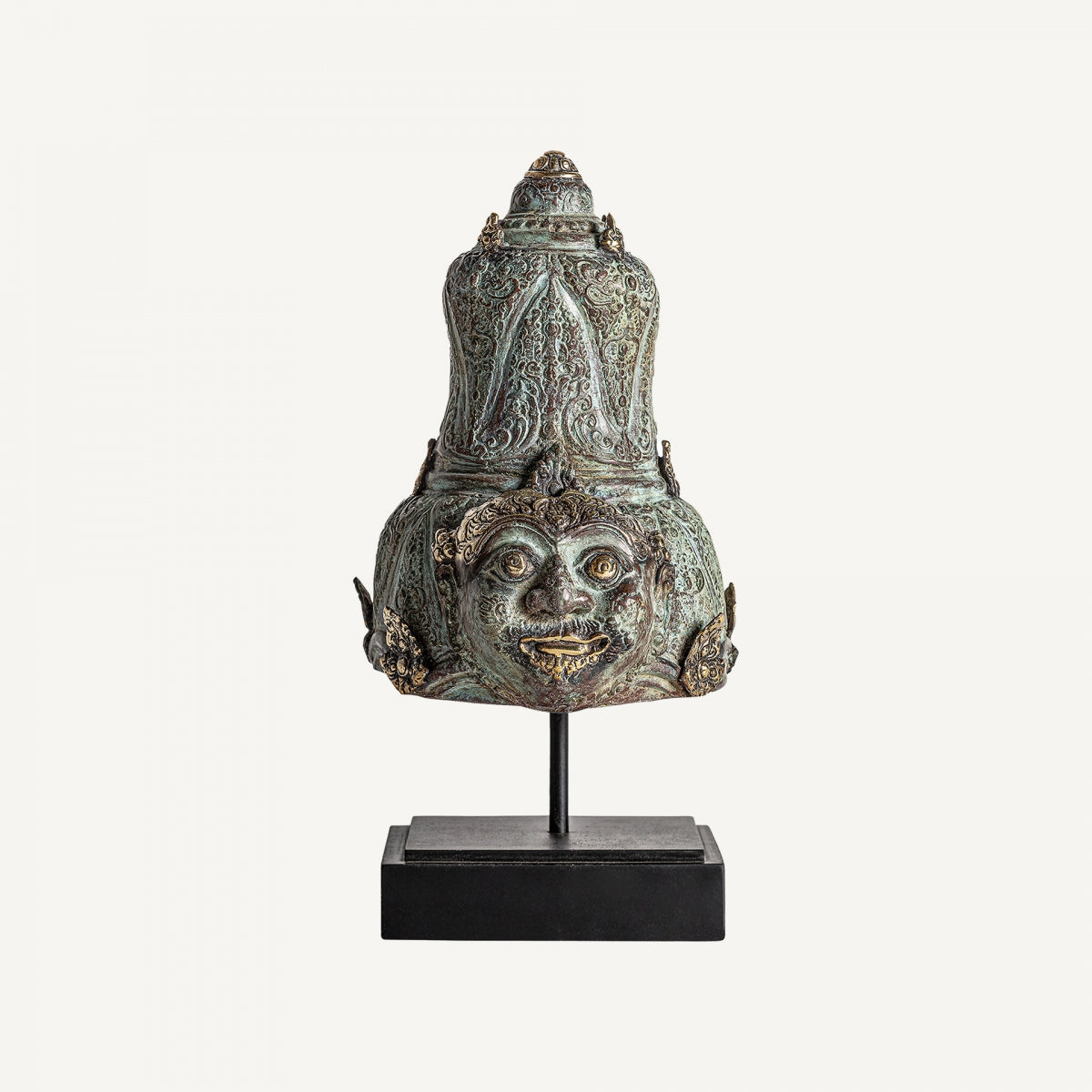 La Figura decorativa oriental en bronce Yasmin presenta un rostro con ojos y dientes prominentes, acabado envejecido y detalles ornamentales. Está montado sobre una base negra y destaca sobre un fondo sencillo.