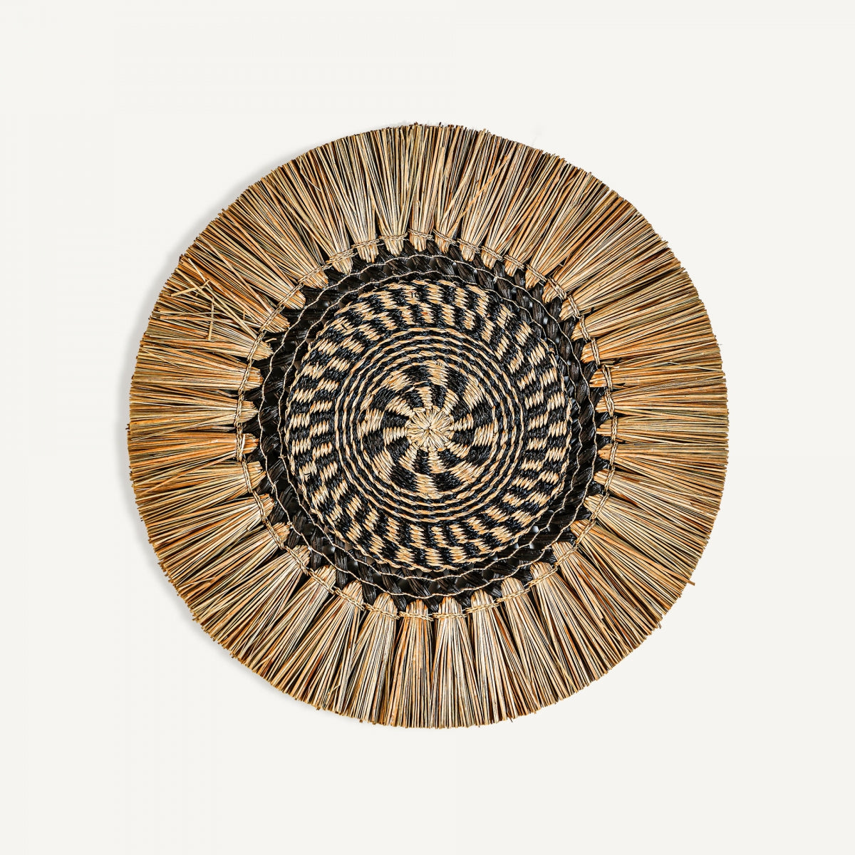 El Panel decorativo en rafia Boho Iris es una bandeja redonda tejida con un diseño geométrico central negro y natural, enmarcado por paja orientada hacia afuera, ideal para agregar un toque boho a tu espacio.