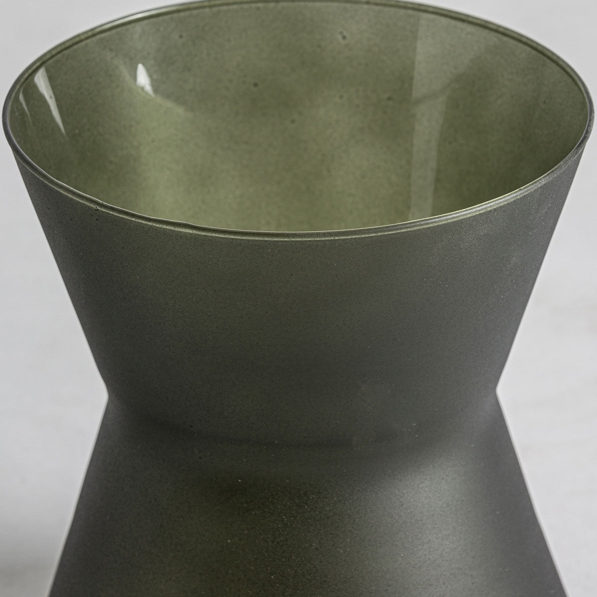 Un primer plano del jarrón de vidrio verde Nehoiu S, con forma de reloj de arena, amplia abertura y acabado verde oscuro mate, sobre un fondo gris claro, ideal para la decoración contemporánea.
