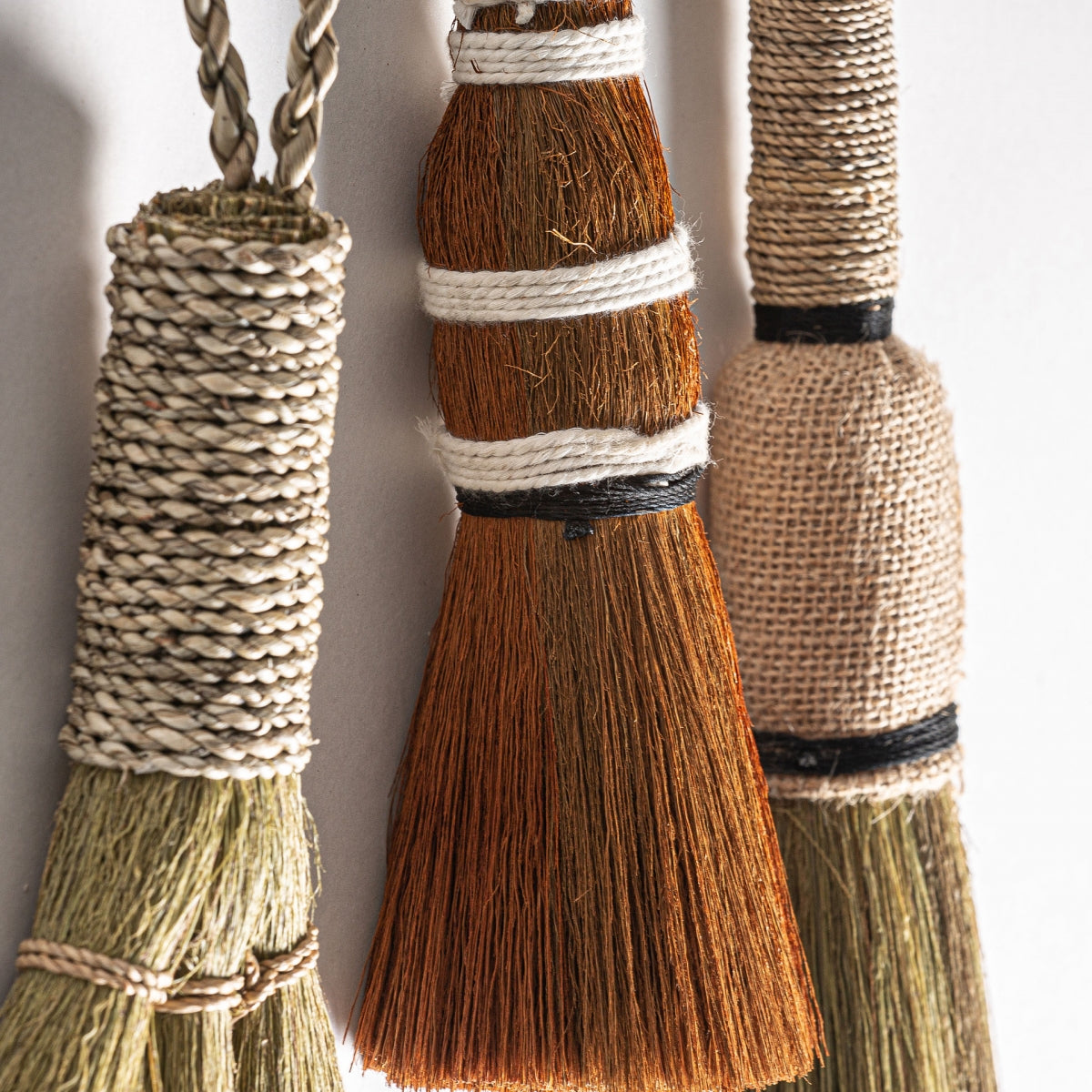 Primer plano de tres Escobillas decorativas en yute con toque étnico S.Wrae, cada una con distintas texturas y colores de fibra natural, colgando verticalmente sobre un fondo claro para un toque de estilo bohemio.