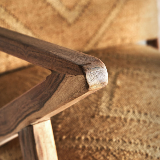 Primer plano del reposabrazos de madera del Sillón de madera y yute Elgin sobre un fondo de tela texturizada y estampada, que desprende un diseño vintage que realza un ambiente rústico.