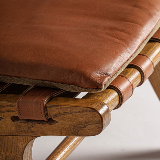 Primer plano de la silla Alford, hecha de piel y madera de fresno, con un cojín de cuero marrón sobre un soporte de correas tejidas. El diseño de inspiración art déco destaca un acabado de madera marrón claro con juntas visibles y correas de cuero.