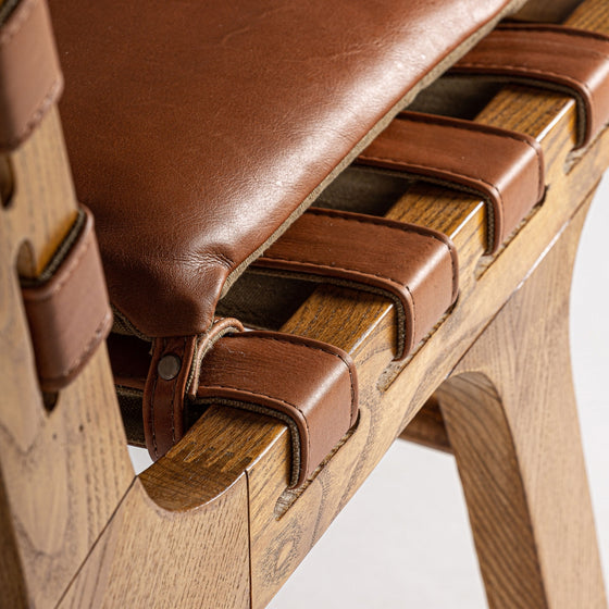 Primer plano del Sillón Alford, con asiento de cuero marrón y estructura de madera de fresno con correas entrelazadas. Esta exquisita pieza refleja el estilo Art Déco con el uso del cuero natural.