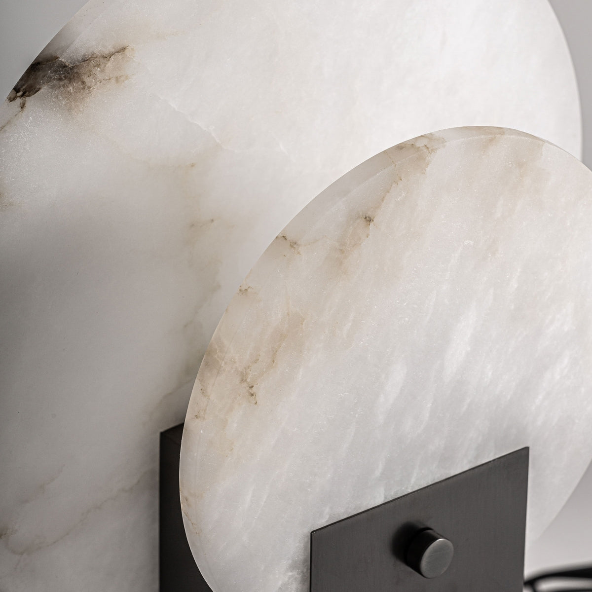 La Lámpara de sobremesa en hierro y piedra Avala presenta dos lasas circulares de mármol blanco con vetas marrones sobre una base de hierro negro, evocando el diseño Art Deco y perfecta como pieza elegante y llamativa para cualquier espacio.