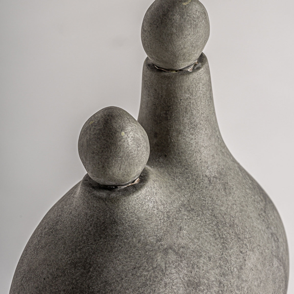 La Figura decorativa en cerámica gris Kedha es una escultura contemporánea con dos formas redondeadas, una mayor y otra menor, cada una coronada por una esfera. Destaca la elegancia de la cerámica gris sobre fondo neutro.