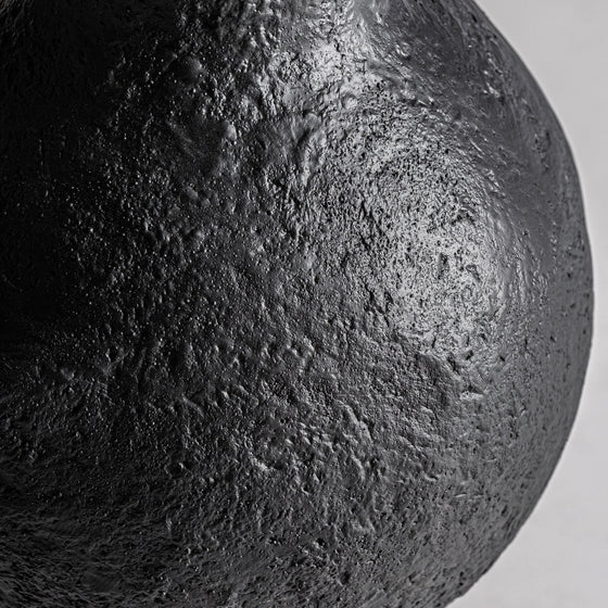 Primer plano del Jarrón de resina negra Riley S, que muestra su superficie oscura y texturizada, similar a una luna o una roca volcánica. Elaborado en resina negra con detalles irregulares y sutilmente iluminado en la parte superior derecha, es una pieza fascinante para la decoración de interiores.