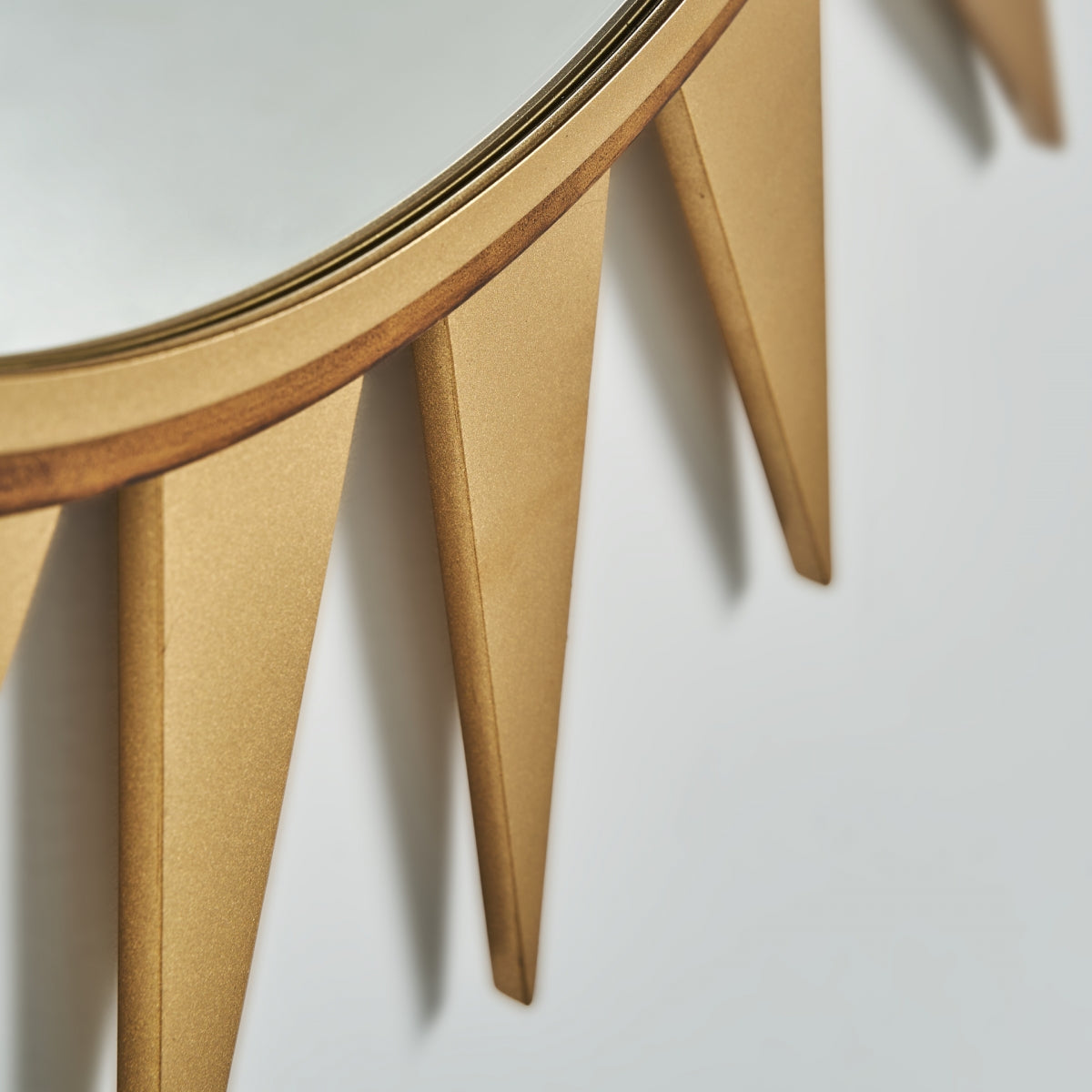 Primer plano de Becky, un espejo redondo de hierro dorado (129x129 cm) con detalles de marco triangular estilo sunburst de estilo art déco, sobre un fondo blanco.