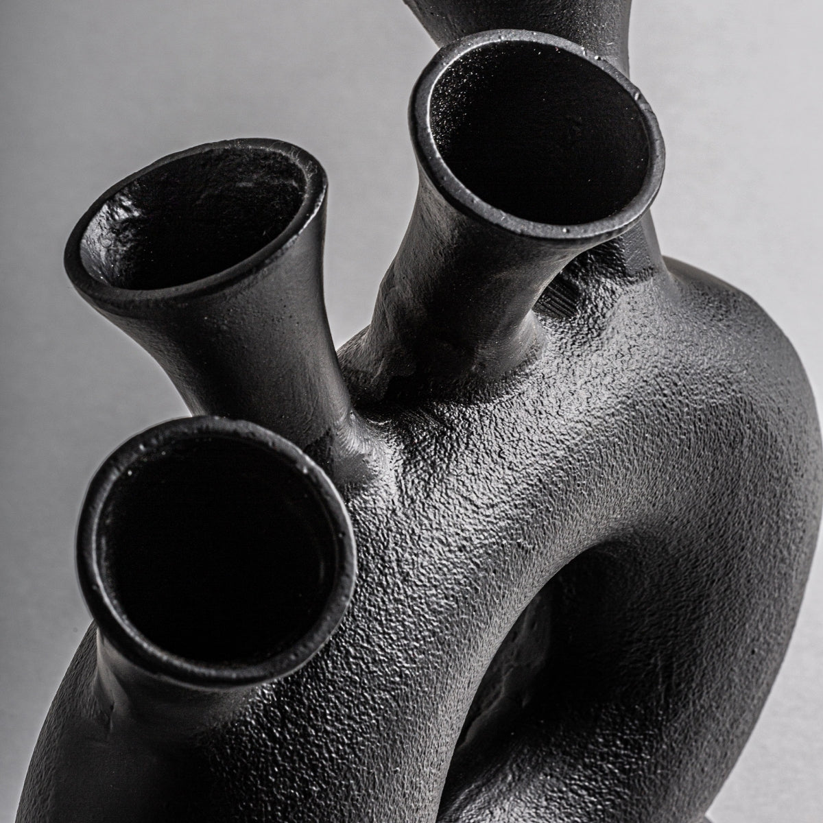 Primer plano de la Figura decorativa en aluminio negro Davet con tres aberturas tubulares y base redondeada, realizada en aluminio negro sobre fondo gris, ideal para los amantes del arte contemporáneo.