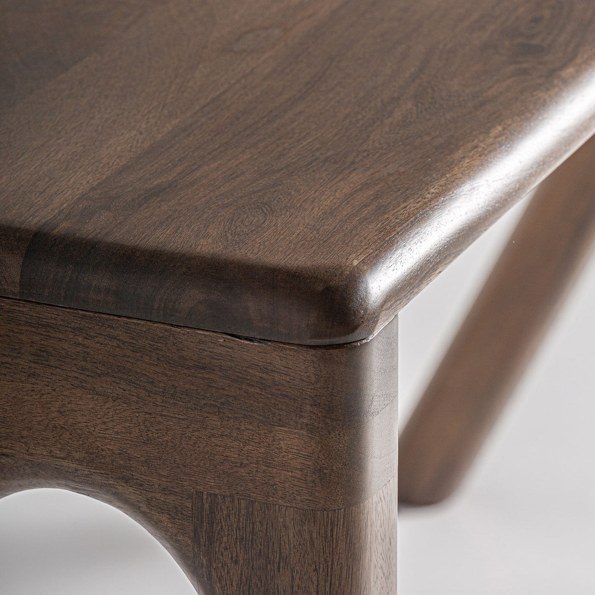 Primer plano de un escritorio en madera de mango Wilsau de esquina, resaltando sus bordes suaves y redondeados y la veta visible de la madera de mango, con parte de una pata en el fondo para un toque contemporáneo.