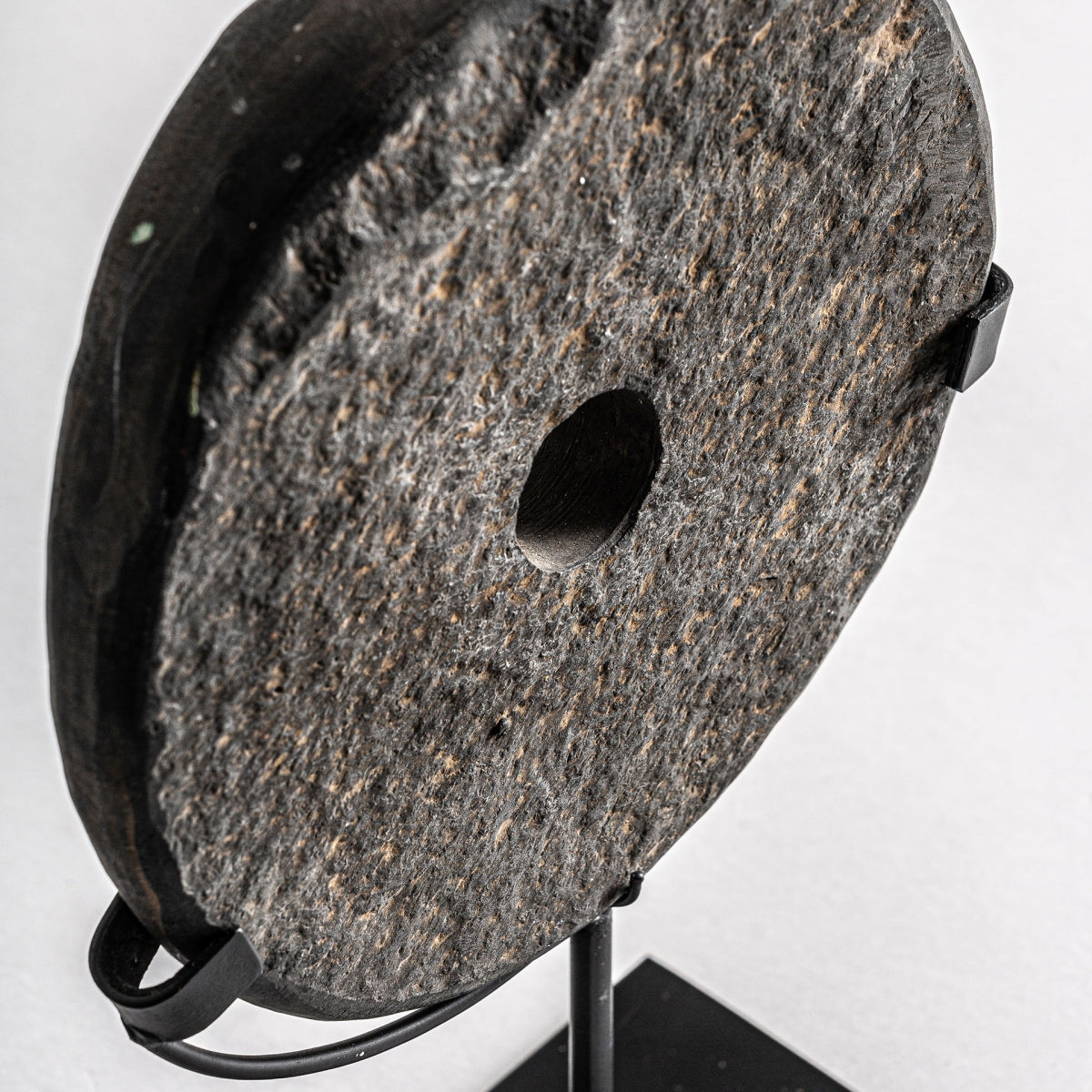La Figura decorativa disco en piedra y hierro Seoane S presenta un disco circular de piedra con orificio central, montado verticalmente en un soporte de hierro negro, perfecto para dar un toque personal a cualquier ambiente.