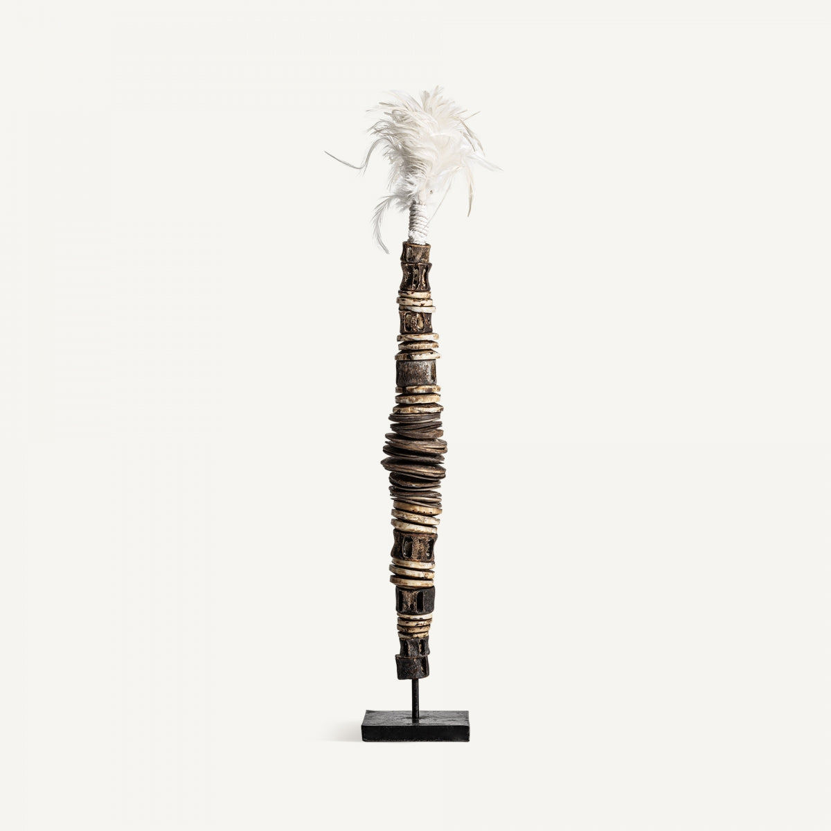 La figura decorativa étnica en hierro y concha Kutubu presenta círculos marrones y beige con plumas blancas sobre soporte negro, de acabado artesanal; ideal para la decoración de interiores.
