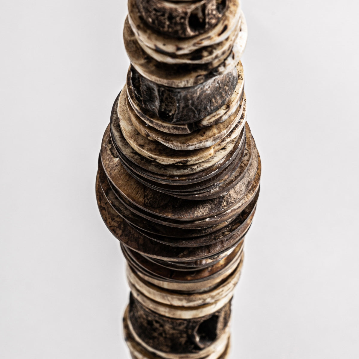 La Figura decorativa étnica en hierro y concha Kutubu presenta una pila vertical de discos irregulares en tonos marrón y beige, con acabado artesanal, vista desde arriba sobre fondo blanco; Ideal para decoración de interiores o como adorno étnico.