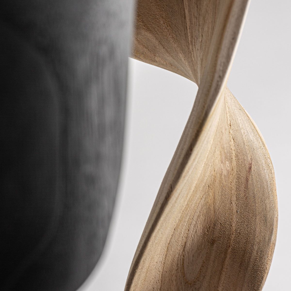 La figura decorativa en madera de teka Blayen, con sus intrincados giros, se asienta elegantemente junto a un objeto curvo oscuro, representando una decoración moderna sobre un fondo liso.
