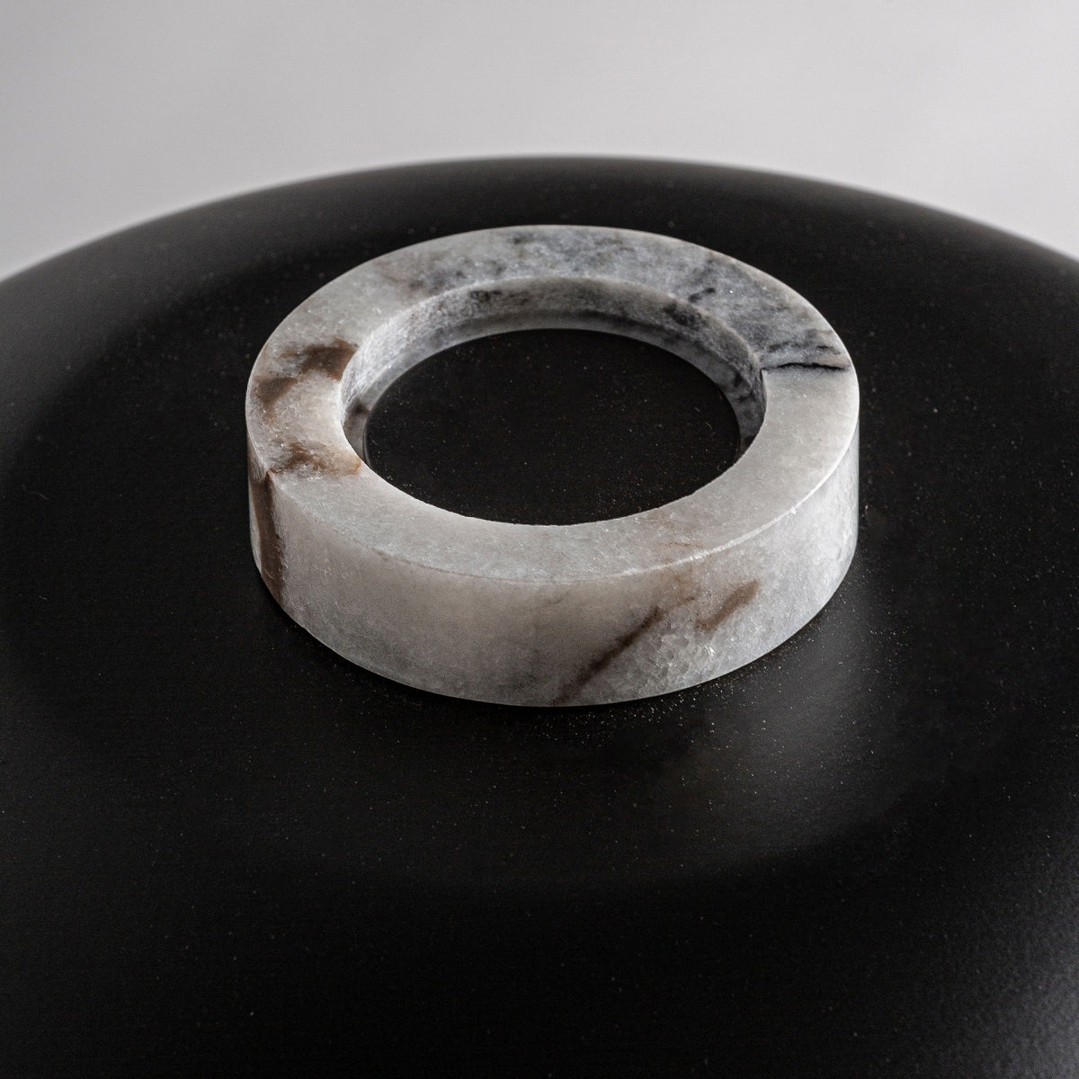 Un anillo redondo de mármol con vetas grises y marrones se asienta sobre una superficie lisa de mármol negro, formando un diseño elegante que recuerda a la Lámpara de mesa en mármol e hierro Midi.
