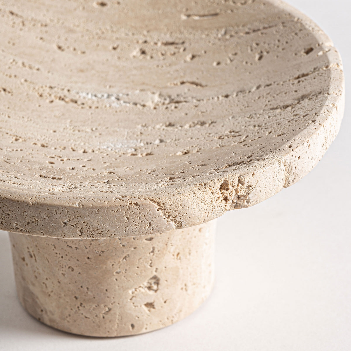 Un primer plano del centro de mesa minimalista en travertino Jasha S, de color beige, redondo, con base cilíndrica y textura natural, perfecto para un estilo de diseño moderno.