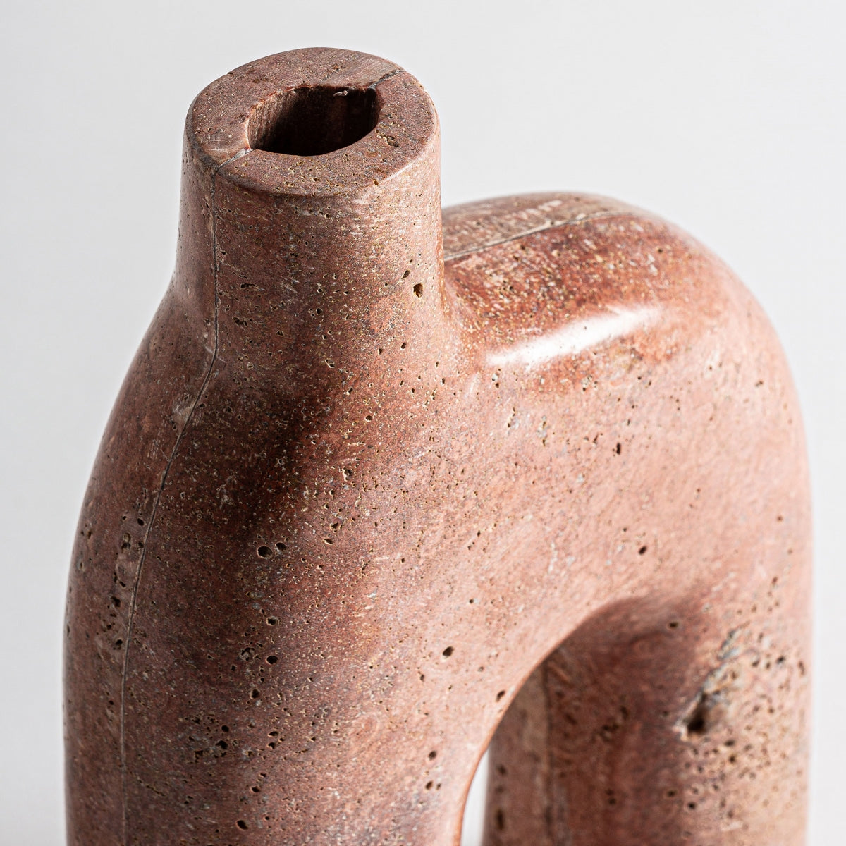 Primer plano de un jarrón de mármol rosa Kleise, liso, de color marrón rojizo, con la parte superior hueca y forma arqueada, que muestra una superficie texturizada y ligeramente porosa inspirada en el diseño art déco.