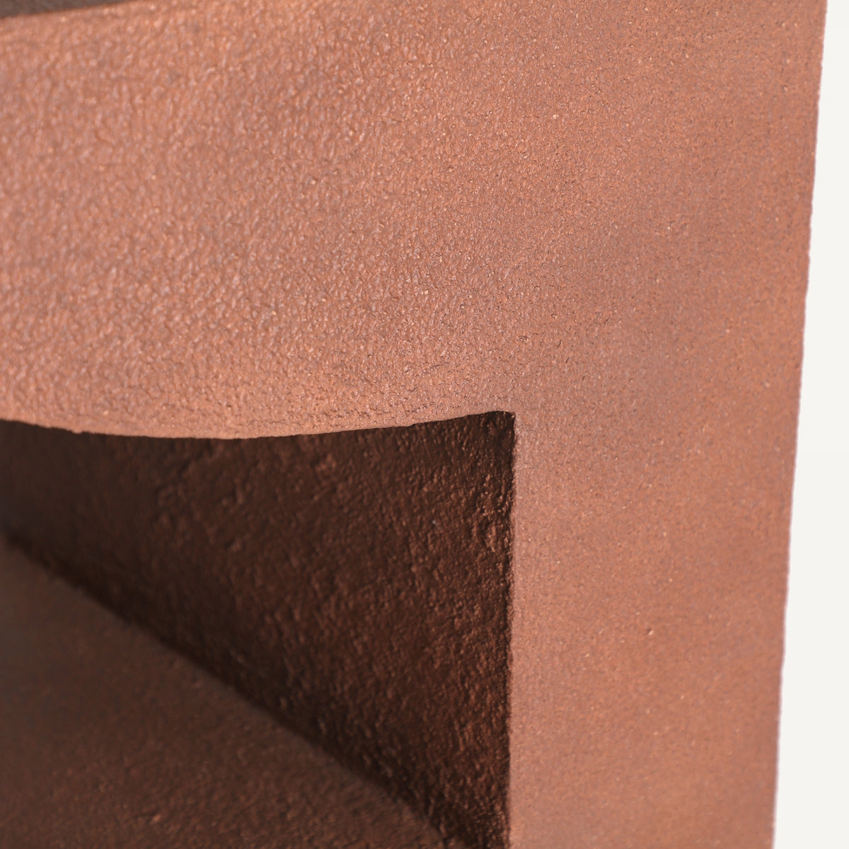 Primer plano de la esquina de la Mesa auxiliar de cemento y madera Steinach, una estructura geométrica de color óxido con ángulos agudos y una sombra triangular insertada, personificación del diseño contemporáneo.