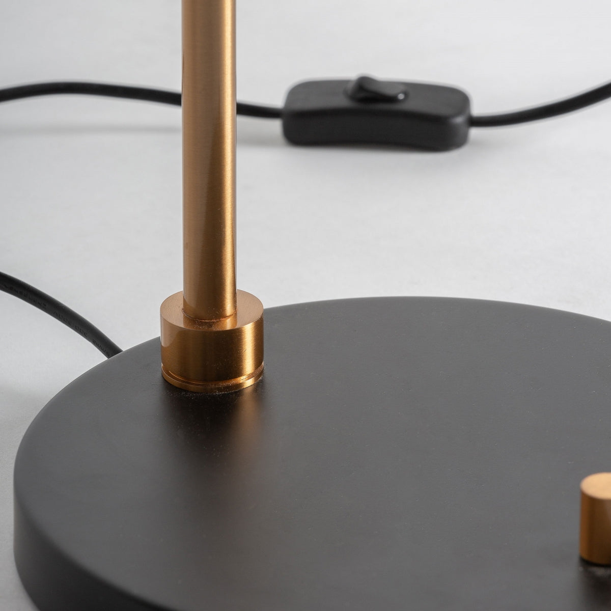 Primer plano de la Lámpara de sobremesa en bronce Eterra, que presenta una base circular negra, una varilla vertical de bronce dorado de alta calidad y un cable de alimentación negro con un interruptor en línea sobre una superficie gris claro, ideal para decoración de interiores.