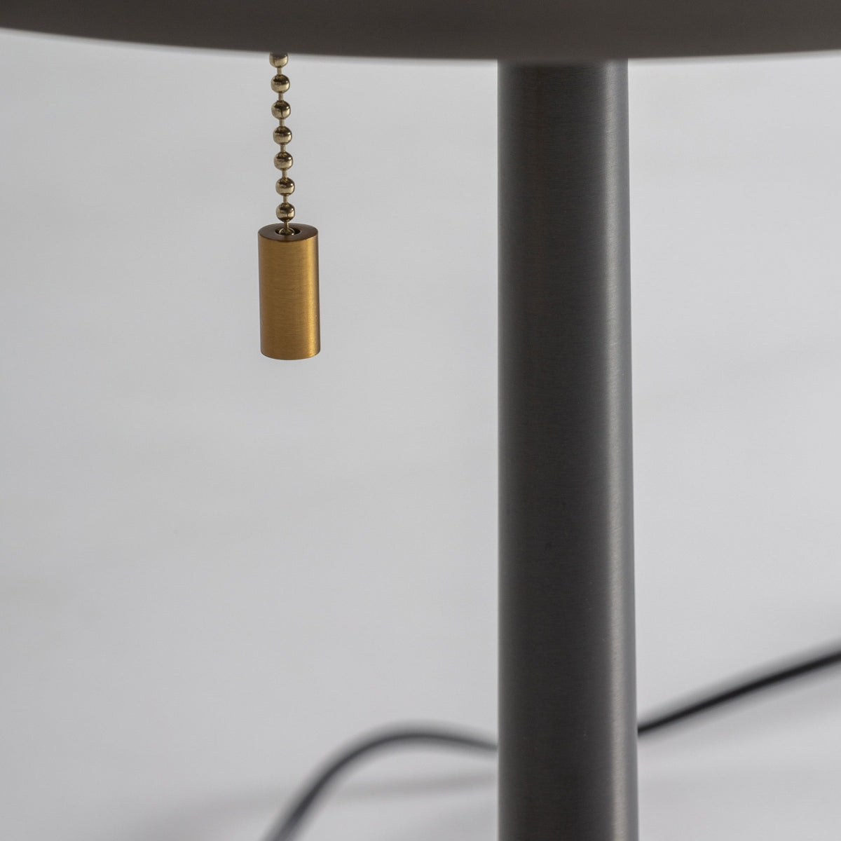 Primer plano de la Lámpara de sobremesa en bronce y dorado Nobia, que cuenta con un soporte metálico y un interruptor de cadena dorado en estilo art decó; un cable negro aparece en el fondo, perfecto para una iluminación ambiental.