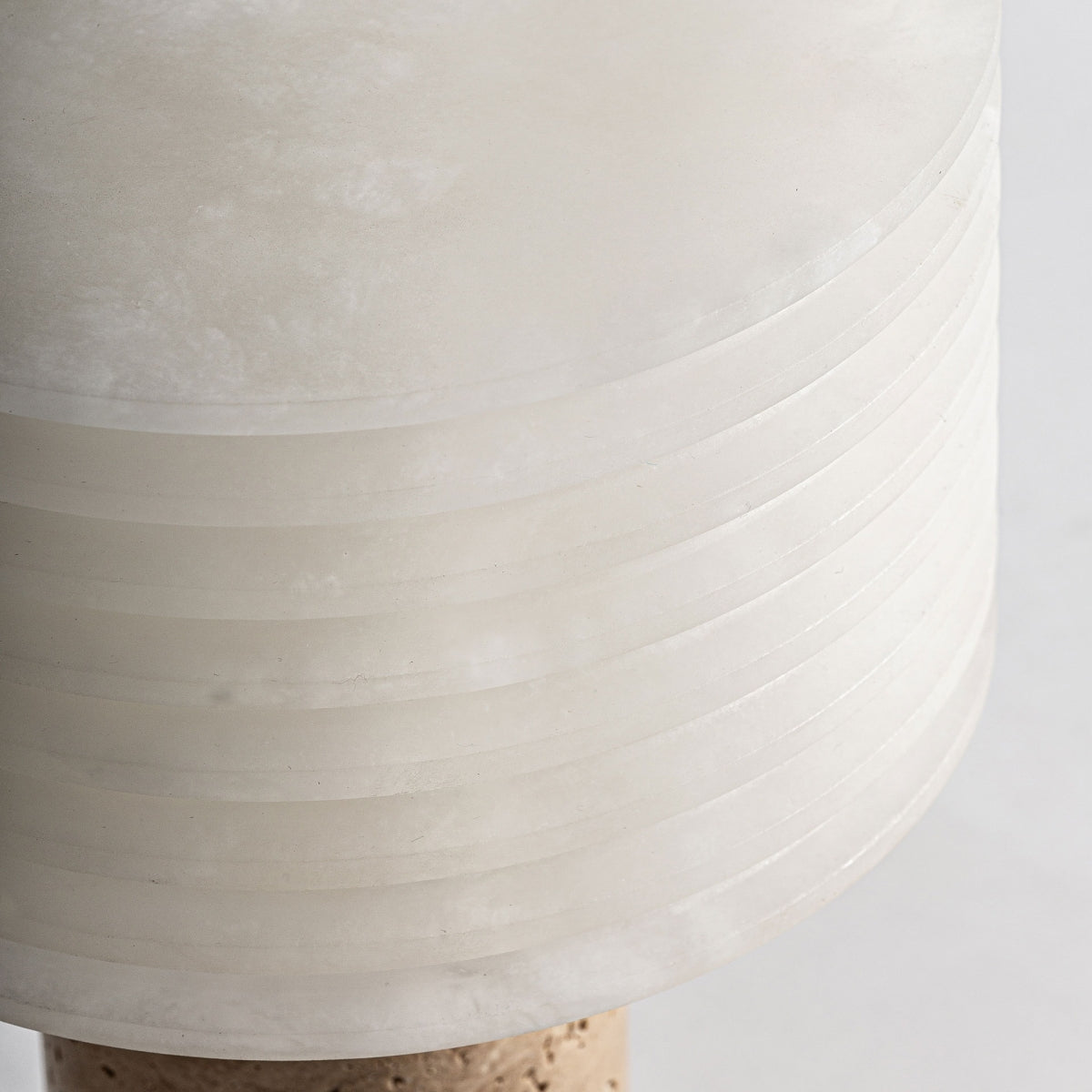 Primer plano de la Lámpara de mesa en cerámica y mármol T-Marb, de diseño art déco con pantalla cilíndrica translúcida blanca y ranuras horizontales, colocada sobre una base de color claro.