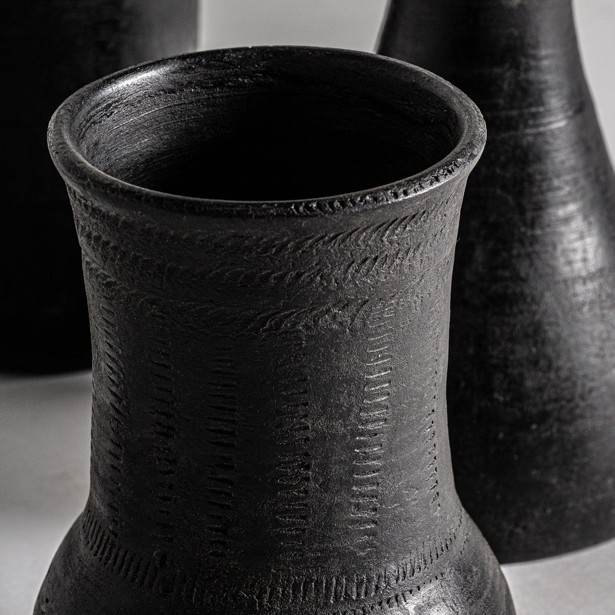 Primer plano de Jarrones de terracota negro Noir (3 piezas) con patrones texturizados y grabados; Dos vasijas similares de estilo moderno son parcialmente visibles al fondo, ideales para una decoración única.