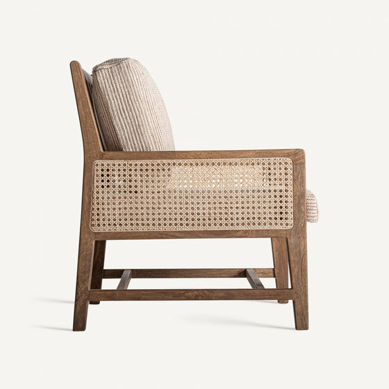 Una vista lateral del Sillón en madera de mango y mimbre Romarand muestra sus detalles de caña tejida y cojines texturizados, que exudan elegancia clásica contra un fondo blanco liso.
