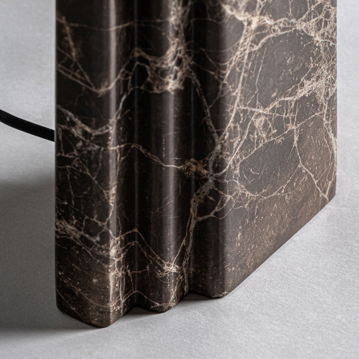 Primer plano de la Lámpara de mesa en mármol marrón y detalles dorados Artis con vetas blancas, cantos estriados y un cable negro a la izquierda, añadiendo un toque elegante y sofisticado al reposar sobre una superficie gris claro.