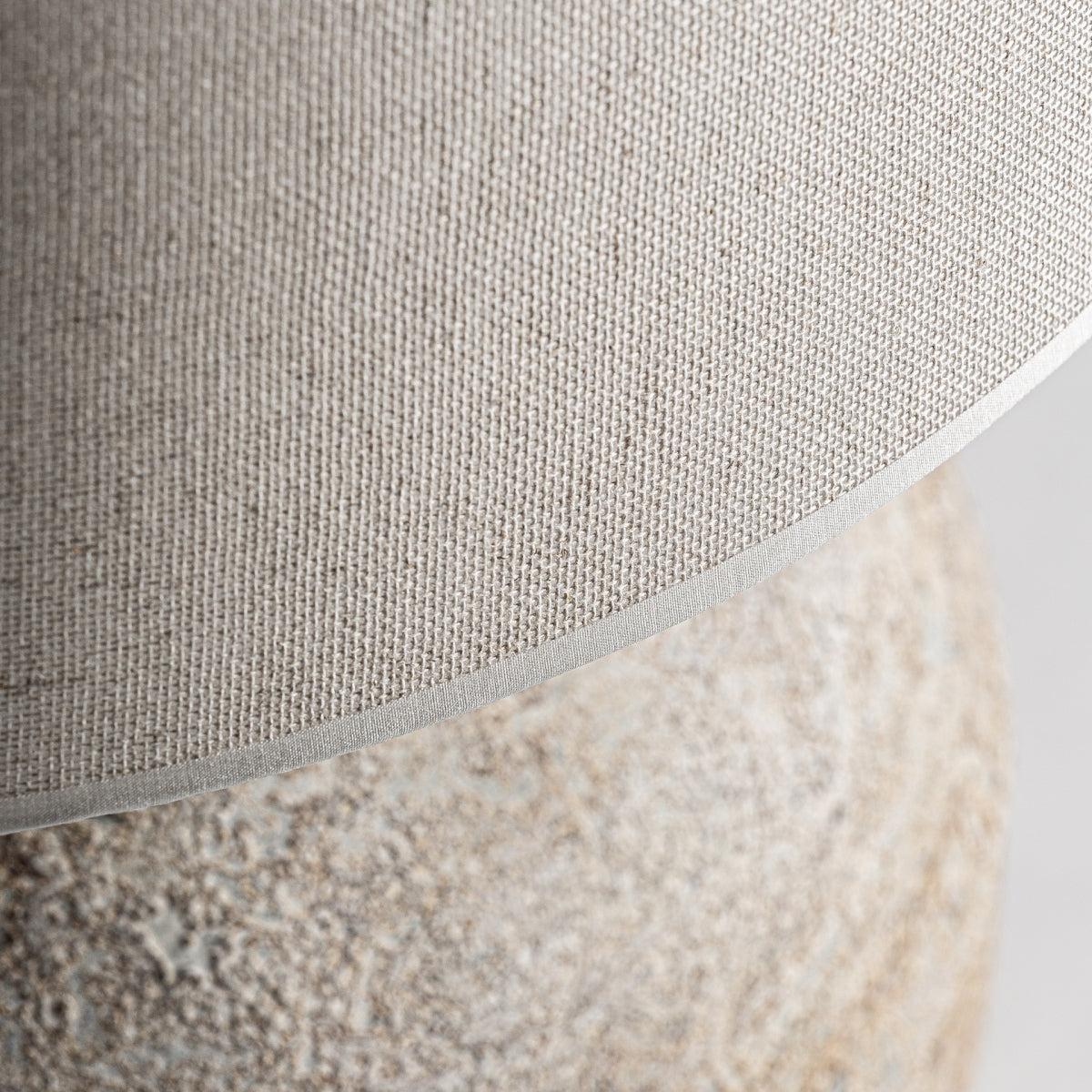 Primer plano de la pantalla de algodón beige texturizado de la Lámpara de sobremesa en cerámica y algodón Provenza, con su base de cerámica similar a una piedra parcialmente visible, que evoca el encanto del estilo provenzal.