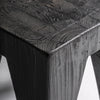 Primer plano del esquinero Taburete bajo en madera reciclada negra Bruschal, destacando su veta texturizada, elaborada a partir de madera de pino reciclado, ofreciendo un diseño versátil que complementa cualquier espacio.
