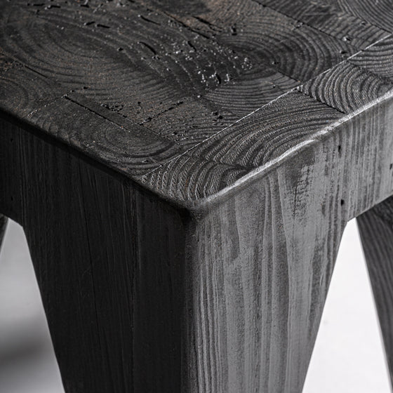 Primer plano del esquinero Taburete bajo en madera reciclada negra Bruschal, destacando su veta texturizada, elaborada a partir de madera de pino reciclado, ofreciendo un diseño versátil que complementa cualquier espacio.