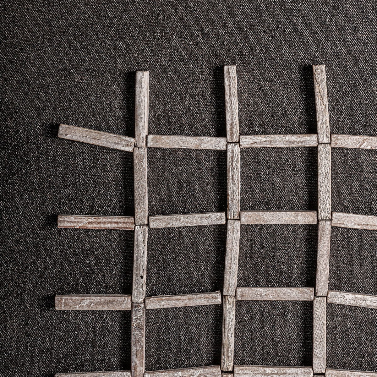 Las varillas de madera del Panel decorativo de madera reciclada tonos oscuros Siegen están dispuestas en cuadrícula sobre tela oscura texturizada. Algunas están desalineadas, lo que le da a esta pieza de decoración sostenible un encanto irregular y único.