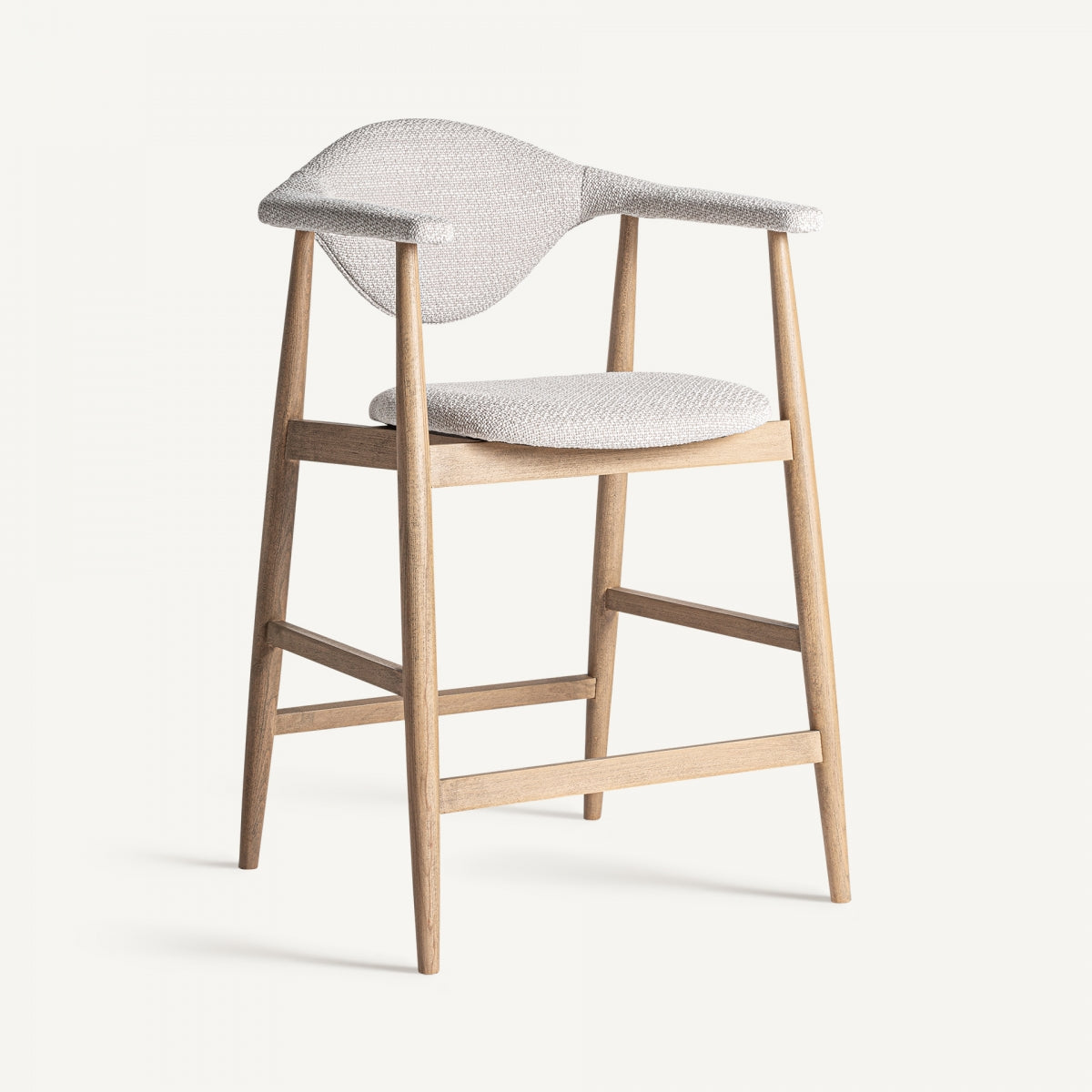 El Taburete de madera de haya y tapizado Dilbeek presenta un tapizado de tela beige claro en el asiento y el respaldo, reposabrazos en ángulo y cuatro patas. Representa un diseño funcional con líneas elegantes que reflejan la fabricación artesanal, ofreciendo una combinación de estilo atemporal y utilidad moderna.
