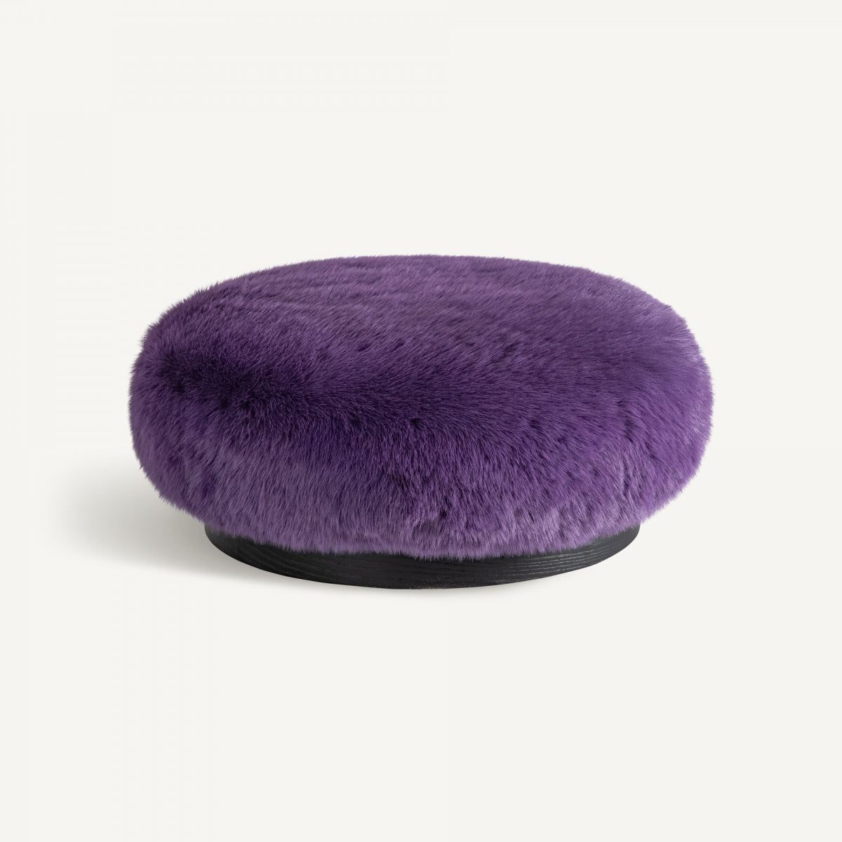 El Puff redondo tapizado en peluche Tropez es un puf redondo de felpa de color morado con una textura peluda que reposa elegantemente sobre una base oscura y resalta su diseño artesanal sobre un fondo liso.