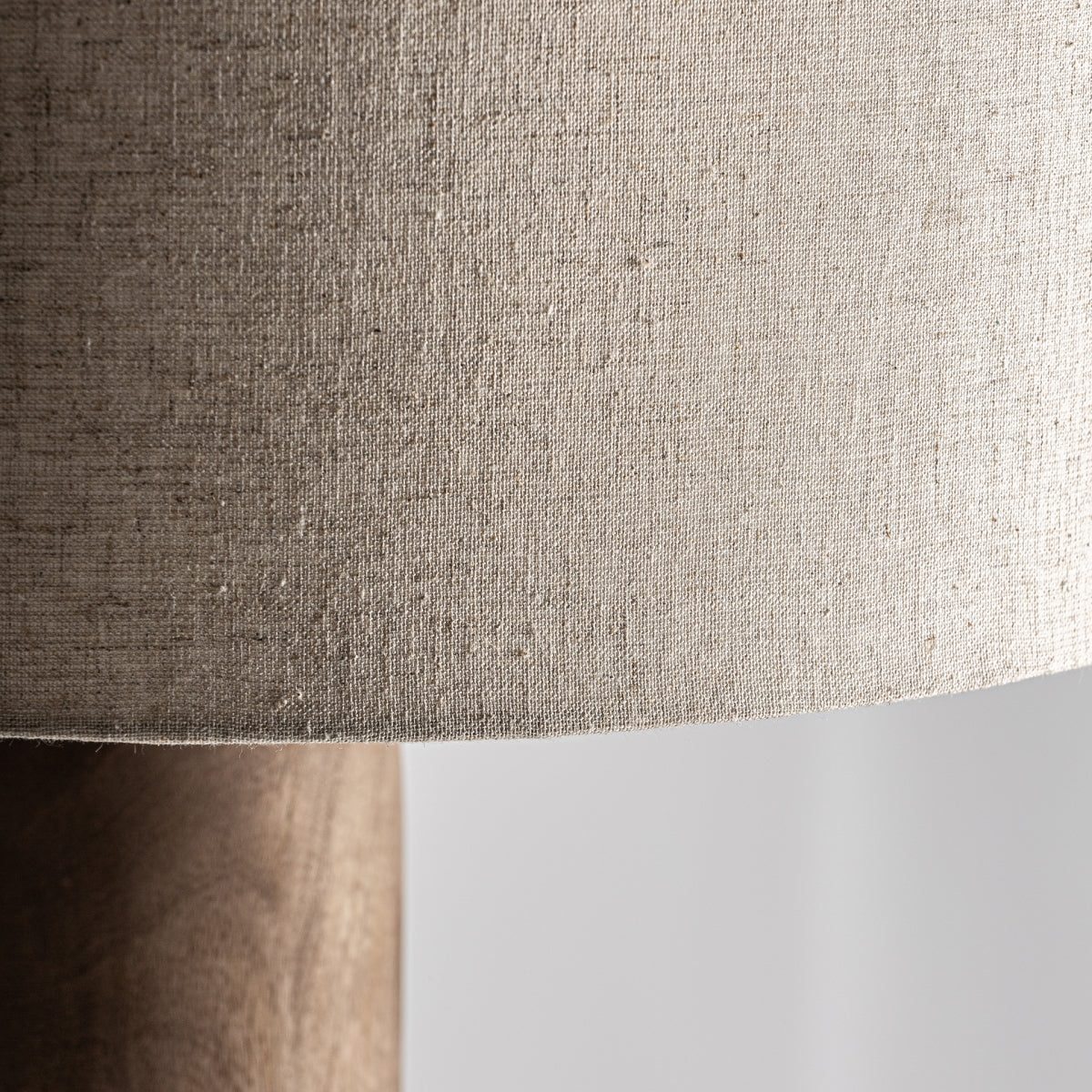Primer plano de una pantalla de tela beige con superficie texturizada, unida a la elegante Lámpara de sobremesa en madera de mango Cogne S, mostrando su diseño elegante sobre un fondo claro.