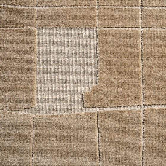 La Alfombra en viscosa Ivana (290x200 cm) presenta un diseño geométrico beige, con una loseta central más clara rodeada parcialmente por tonos oscuros. Las costuras y textura realzan la alta calidad de esta alfombra.