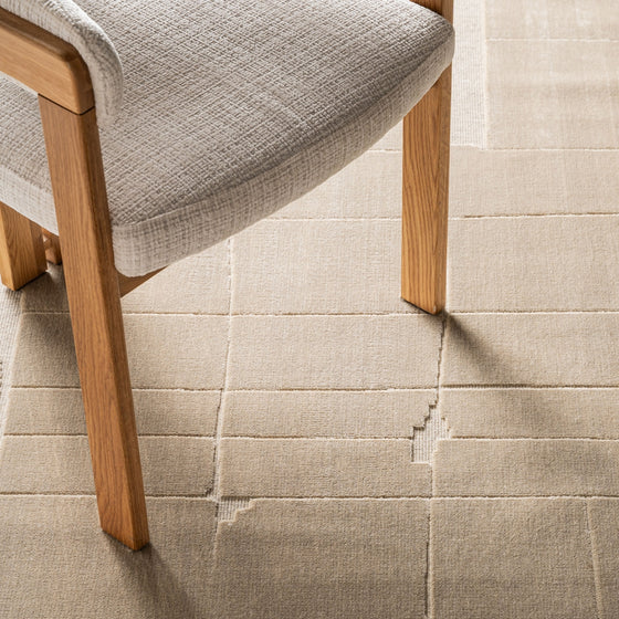 Un primer plano de una silla de madera clara con tapizado texturizado beige sobre la Alfombra en viscosa (290x200 cm) Ivana, que presenta un diseño geométrico beige de alta calidad.
