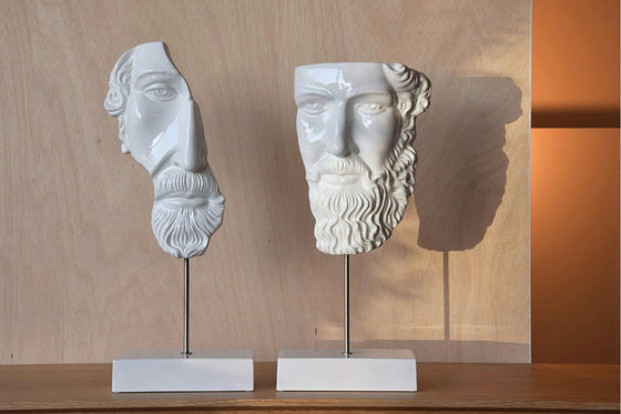 Dos bustos escultóricos de cerámica blanca fragmentados, denominados "Figura decorativa de cerámica blanca griega", se yerguen elegantemente sobre una superficie de madera, encarnando un estilo artístico clásico reinventado. La cálida iluminación proyecta sombras intrigantes en el fondo.