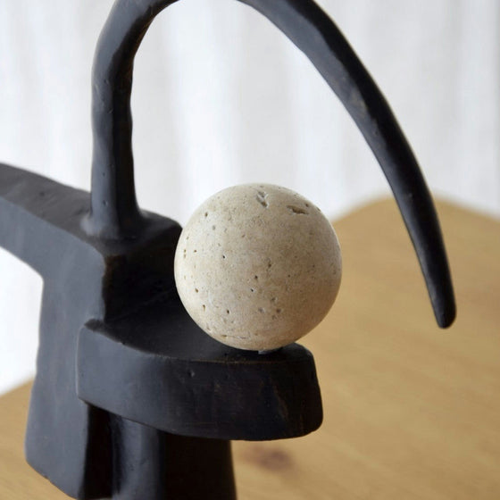 La Figura Decorativa en Metal y Travertino Rula presenta una esfera de piedra texturizada de exquisito mármol travertino sobre una escultura abstracta de metal negro con un brazo curvo, que descansa elegantemente sobre una superficie de madera.