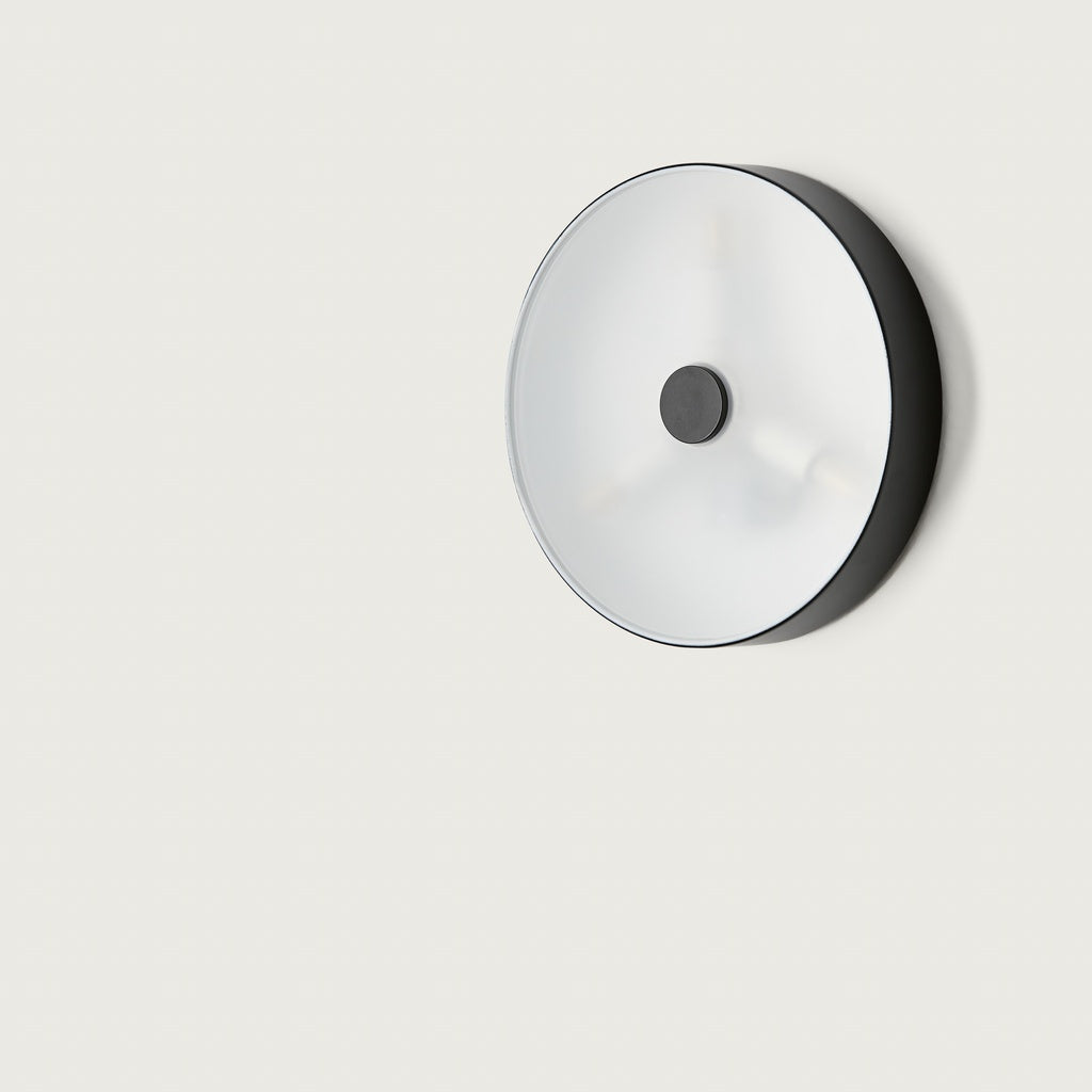 El aplique de pared Ambor es un accesorio redondo y minimalista con borde negro, cuya cubierta blanca esmerilada proporciona luz cálida y envolvente sobre un fondo blanco.