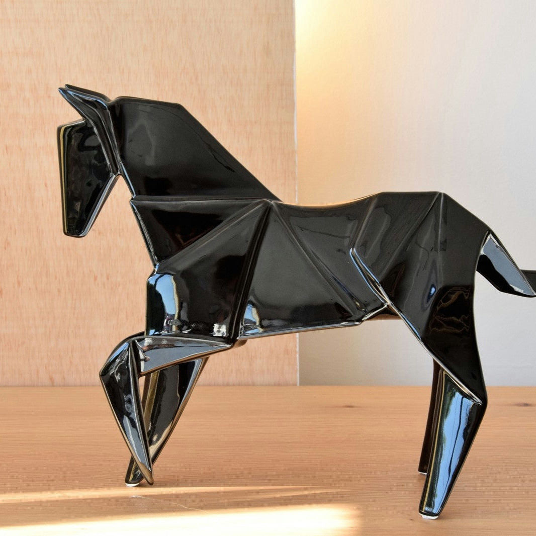 La Figura decorativa en cerámica Negro brillo Caballo Origami muestra un caballo de cerámica negra en estilo origami. Sobre un fondo de madera clara y una superficie de madera, encarna la estética contemporánea con su diseño elegante y moderno.