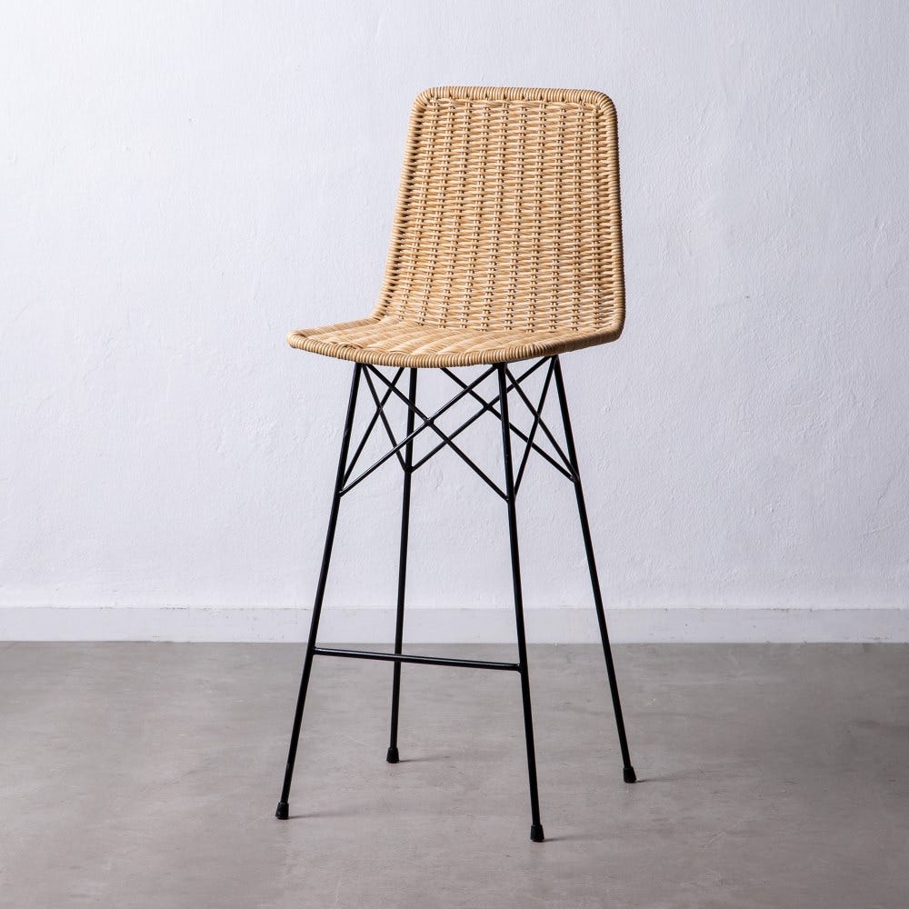 El Taburete alto en ratán sintético y metal Priro, con un asiento de mimbre sobre patas de metal negro, se destaca elegantemente contra una pared blanca lisa sobre un piso de concreto, lo que refleja el elegante espíritu de diseño de CASAPATIO.