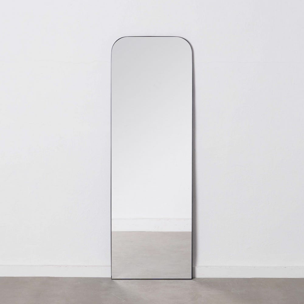 El espejo vestidor en metal negro Orbis, con un diseño de rectángulo alto con bordes superiores redondeados y un elegante marco de metal negro, se apoya contra una pared blanca sobre un piso gris claro, incorporando un diseño moderno.