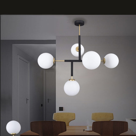 La lámpara de techo de acero y vidrio T-Ball 5, con luces blancas tipo globo y un elegante marco negro, cuelga elegantemente sobre una mesa de comedor de madera con sillas claras. Las paredes y la puerta grises del fondo realzan su estilo contemporáneo.