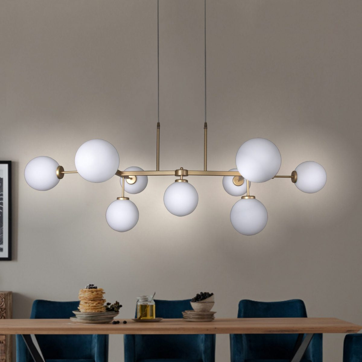 La lámpara de techo en acero y oro Vela 7 cuelga sobre una mesa de comedor con platos, velas y panqueques, rodeada por sillas azules y un cuadro en la pared, resplandeciendo con un toque sutil en color oro.