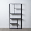 La Estantería Metálica Industrial Negro Square (Alta), una estantería alta de metal negro con un diseño asimétrico, ejemplifica el almacenamiento eficiente contra una pared blanca lisa sobre un piso gris.