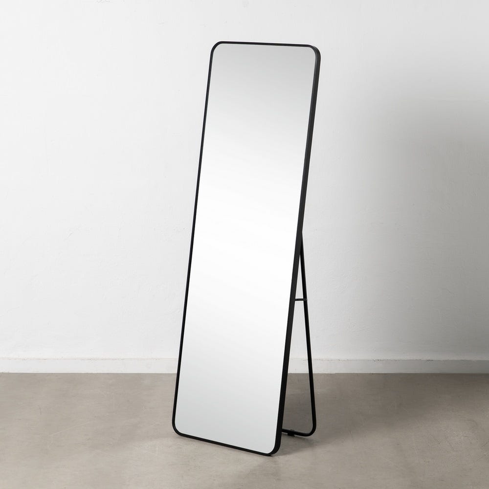 El Espejo Vestidor Aluminio Negro Ortan, un espejo alto y rectangular con marco de aluminio negro, se encuentra sobre un piso gris contra una pared blanca, ofreciendo un diseño versátil que complementa cualquier habitación.