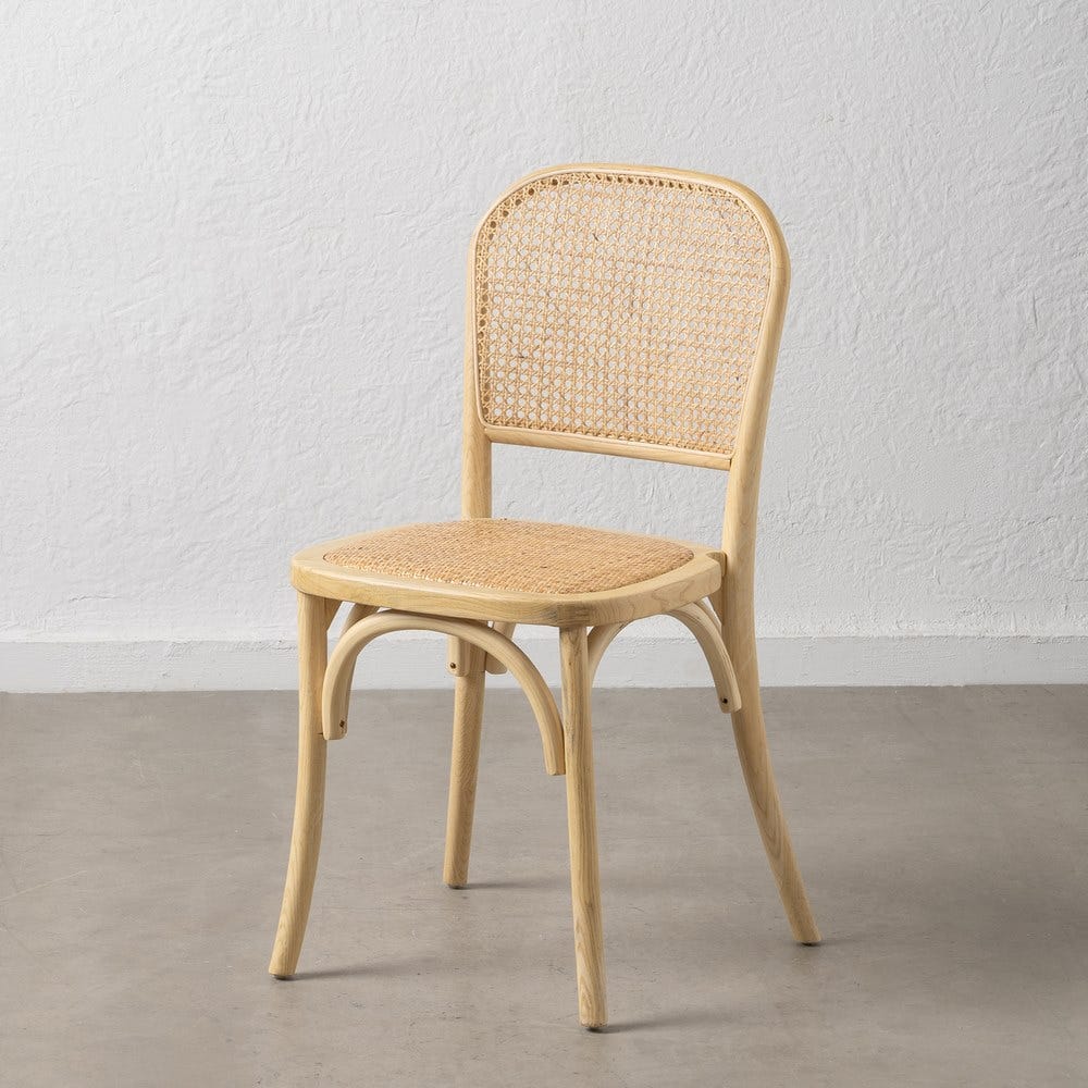 La silla de comedor en madera y ratán Norma, con marco de madera y respaldo y asiento de ratán tejido, reposa elegantemente sobre un piso de concreto gris; su diseño rústico realza la pared blanca lisa detrás de ella.