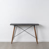 La consola en DM y metal Oslo presenta un diseño minimalista con una elegante tapa rectangular negra, patas de madera y barras transversales de metal. Fabricada con materiales de alta calidad, se destaca elegantemente contra una pared blanca sobre un piso gris claro, lo que encarna un diseño moderno.
