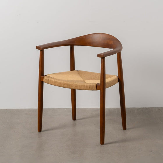 La Silla de Comedor en Madera de Haya Corda, con respaldo curvo y asiento tejido de Cuerda de Papel, está elegantemente colocada sobre el piso de concreto gris.