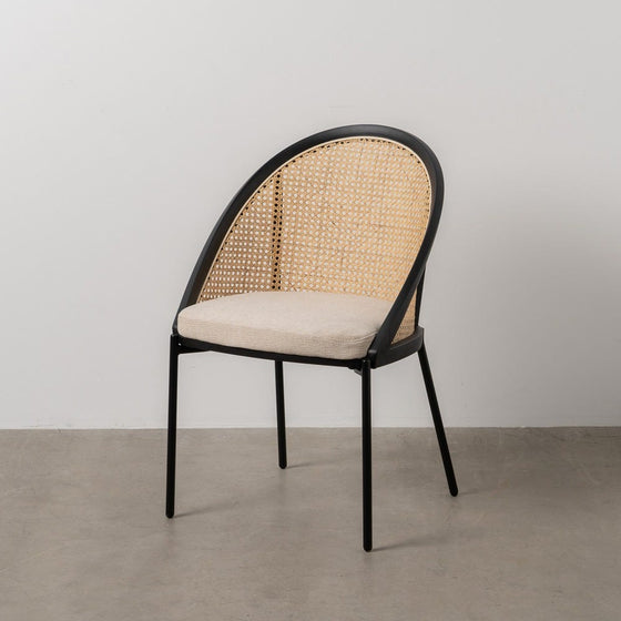 La Silla de Comedor en Acero y Plástico Ratán Alfa, con una estructura de metal negro, respaldo de ratán tejido y asiento acolchado de color beige sobre un piso de concreto liso, encarna el estilo contemporáneo con sutiles toques de diseño industrial.